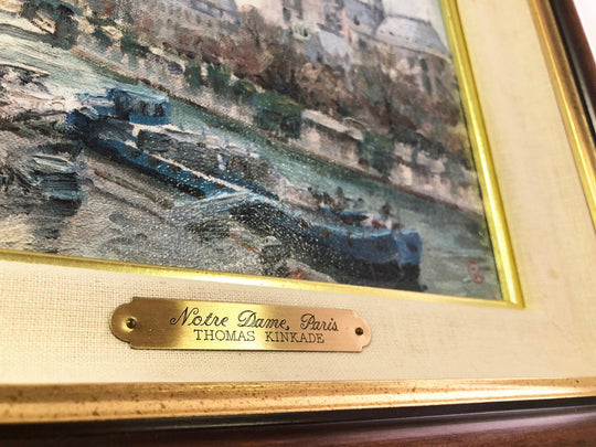 Thomas Kinkade painting of Notre Dame Cathedral in Paris with the River Seine and a flower-lined sidewalk.
